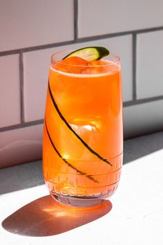 an orange drink with a slice of cucumber on the rim in front of a brick wall