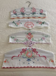 four pieces of embroidered clothing hang on a bed with white linens and pink flowers