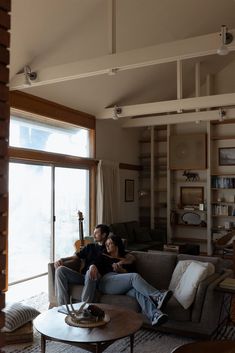 two people sitting on a couch in a living room next to a large open window