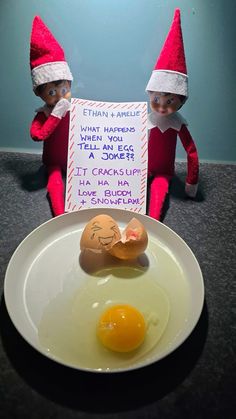 two elfs are sitting next to an egg on a plate with a sign in front of them