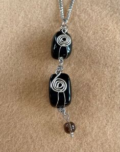 This pendant necklace has been made using 2 Obsidian gemstone, drilled cabochons, which have been decoratively wrapped with silver plated wire, embellished with a coiled, silver plated wire spiral and linked together to form a double length pendant. The base of the pendant has a smoky quartz pendant bead and the whole pendant is hung on a chunky, 3mm flat curb chain, with a lobster claw clasp. Pendant total length: 8cm Pendant width: 2.5cm Necklace length: 16"(40.5cm) Obsidian gemstones: approxi Spiral Wire Wrapped Sterling Silver Necklace, Silver Obsidian Necklaces, Silver Obsidian Necklace With Natural Stones, Adjustable Silver Obsidian Necklace, Silver Obsidian Jewelry With Gemstone Beads, Elegant Black Spiral Jewelry, Black Agate Wire Wrapped Jewelry, Spiral Black Necklace As A Gift, Black Spiral Necklace As A Gift