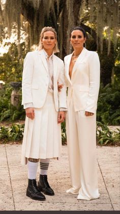 two women dressed in white standing next to each other on a sidewalk with trees behind them