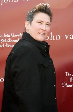 a woman standing in front of a red wall wearing a black coat and smiling at the camera