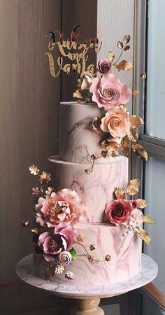 a three tiered wedding cake with pink flowers and gold lettering on top, sitting in front of a window