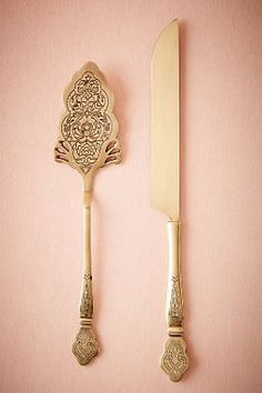 two golden utensils are next to each other on a pink surface, one is holding a knife and the other has a gold handle