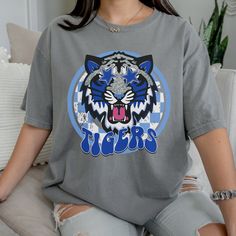 Get this popular new Tigers Mascot shirt and show off your school spirit in style!  Each shirt is printed in-house by me using high-quality materials on a Comfort Colors shirt, known for its exceptional comfort, style, and durability. Even the glitter is printed! No need to worry about peeling glitter vinyl, or messy glitter flakes. Available in unisex sizes S to 3X, and youth sizes, we've got you covered regardless of your preferred fit. The Preppy Tigers Shirt features a classic crew neck and short sleeves, making it suitable for year-round wear. The flattering silhouette ensures a comfortable and stylish fit for both men and women. Please note this listing is ONLY for comfort colors grey Note: As each shirt is individually printed, slight variations in color and placement may occur, add School Spirit T-shirt With Graphic Print, School Spirit Shirt With Screen Print For Game Day, School Spirit Graphic Print Top, School Spirit Game Day Shirt With Screen Print, Collegiate Graphic Print Tops For School, Casual School Shirt With Sublimation Print, Casual College Shirt With Sublimation Print, College Style Graphic T-shirt For School, Graphic Print College Style Tops For School