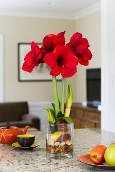 red flowers in a glass vase on a table with other fruit and vegetables around it