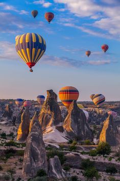 many hot air balloons flying in the sky over some rocks and trees, with blue skies above