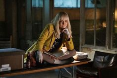 a woman sitting on top of a wooden table next to a window holding a book