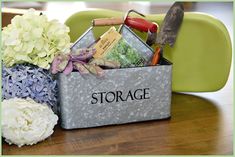 a storage container filled with flowers and gardening tools on top of a wooden table next to a green chair