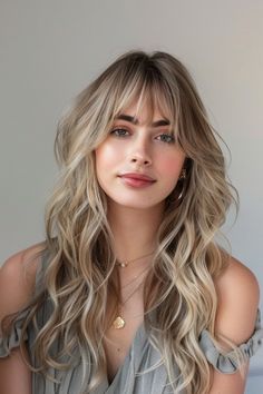 Woman with long wavy blonde hair and bangs, wearing gold jewelry and a gray ruffled top, looking at the camera. Wavy Ash Blonde Hair, Blonde Hair For Light Olive Skin, Highlights For Ash Blonde Hair, Ash Roots Blonde Hair, Ash Blonde Hair Highlights, Ash Blonde With Highlights, Blonde Hair Ashy, Winter Hair Colour For Blondes, Ash Blonde Hair Color Ideas