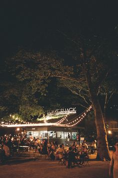 people sitting at tables in front of a building with lights strung from the roof and trees