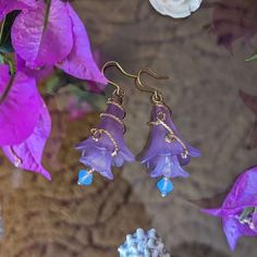 Purple flowers have a subtly asymmetric accent of gold wire. The lower flower and blue bead jangle a little as the wearer moves their head. Whimsical Gold Flower Earrings For Party, Gold Whimsical Flower-shaped Earrings, Delicate Blue Flower Earrings With Ear Wire, Whimsical Blue Flower Earrings, Whimsical Gold Flower Earrings, Whimsical Gold Flower Earrings For Wedding, Bohemian Gold Flower Earrings Handmade, Flower Statement Earrings, Lucite Jewelry