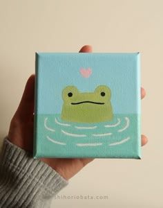 a person holding up a small square painting with a frog on it's face