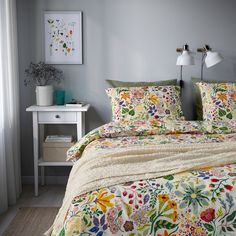 a bed with floral sheets and pillows in a room next to a table, lamp and pictures on the wall