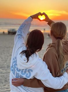 two girls making a heart shape with their hands on the beach as the sun sets