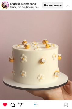 a hand holding a white cake with yellow bees on top and flowers around the edges