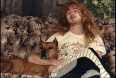 a man with long hair sitting on a couch next to a brown and white dog