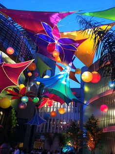 many colorful lights hanging from the ceiling in an indoor area with palm trees and tall buildings