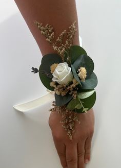 a woman's hand holding a white rose and greenery corsage on her arm