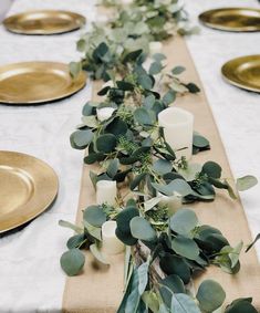 the table is set with gold plates and greenery on it, along with candles