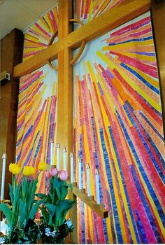 a cross with candles and flowers on it in front of a wall painting that is made from strips of colored paper