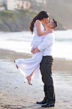 a man holding a woman while standing on top of a beach next to the ocean