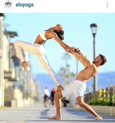 two people are doing yoga on the sidewalk near some buildings and street lights, one man has his leg up in the air while the other man is leaning forward