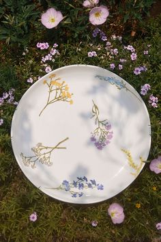 a white plate with flowers on it sitting in the grass next to pink and purple flowers