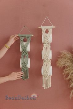 two macrame wall hangings on a pink wall next to a woman's hand