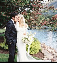 a bride and groom standing next to each other in front of a lake with flowers