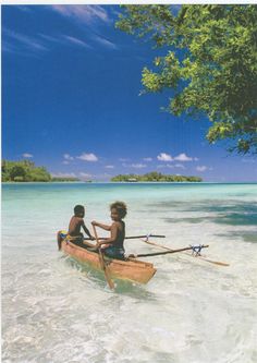 two people in a canoe paddling through shallow water on an island with trees and blue sky