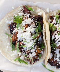 two tacos with meat, cheese and herbs on them sitting on a white plate