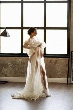 a woman standing in front of a window wearing a white dress with long sleeves and high slits