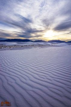 the sun is setting over an expanse of white sand and hills in the distance, with mountains in the distance