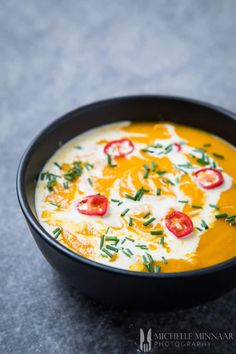 a black bowl filled with yellow soup and topped with cheese, tomatoes and green garnish