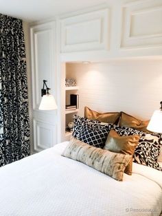 a white bed topped with lots of pillows next to a window covered in black and white curtains