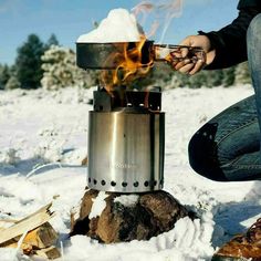 a person sitting in the snow next to a stove with fire coming out of it