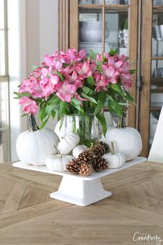 pink flowers and white pumpkins in a glass vase on a tray with pine cones