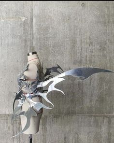 a white mannequin with metal wings on it's head and torso, standing in front of a concrete wall