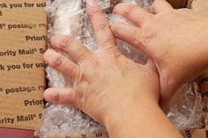 two hands reaching for something out of a box that is wrapped in plastic and cardboard