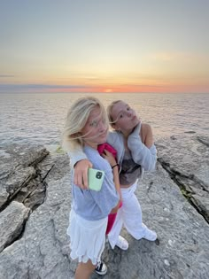 two young women standing on top of a rocky cliff next to the ocean at sunset