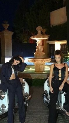 two women standing next to each other in front of some parked cars and a fountain