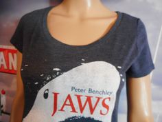 Peter Benchley, Jaws t-shirt. Jaws, the famous novel turned adapted to a screenplay resulting in one of the most famous movies ever made. Fitted tee; curved, fitted for a female's body/ A blue tee featuring graphic and author's name on front. Bright. Fun. Perfect slight fade. Bold yet dainty. Clean. No issues. Ready to wear. Tagless tag states Medium and the measurements are: 23 1/2 inches long (top to bottom), 17 inches across (armpit seam to armpit seam). https://vintagethisexit.etsy.com Jaws is a 1974 novel by American writer Peter Benchley. It tells the story of a great white shark that preys upon a small resort town and the voyage of three men trying to kill it. The novel grew out of Benchley's interest in shark attacks after he learned about the exploits of Montauk, New York shark fi Fitted Crew Neck Shirt With Front Print, Fitted T-shirt With Front Print And Crew Neck, Fitted Crew Neck T-shirt With Front Print, Fitted Graphic Tee For Fan Merchandise, Fitted Graphic Print Tops For Fan Merchandise, Fitted Tops With Graphic Print For Fan Merchandise, Fitted Crew Neck Shirt With Screen Print, Fitted Crew Neck Shirt With Band Merch, Fitted Short Sleeve T-shirt For Fan Merchandise