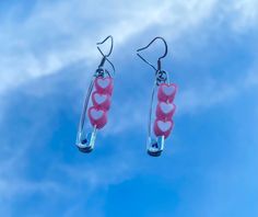 two pink heart shaped earrings hanging from hooks in front of a blue sky with clouds