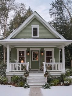 a small green house with white trim and porches in the front yard covered in snow