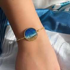 Dive into the depths of style with our ocean and beach-themed bracelet, a perfect accessory for those who adore the sea.  Adjustable up to 20 cm in length, this bracelet features a 22 mm diameter ring adorned with a captivating oceanic and beach view. Crafted with meticulous attention to detail, all metal parts are 24K gold-plated, ensuring both durability and elegance. Handcrafted with resin and resin pigments, each piece is unique, embodying the serene beauty of the ocean. Whether you're a beachcomber or a sailor at heart, this bracelet is sure to make a splash. Packaged with care, it makes for an ideal gift for any ocean lover. Let the waves of style wash over you or surprise a loved one with a piece of the sea. Order now and let the adventure begin! Due to the nature of handmade produc Beachy Sand-colored Jewelry For Gift, Beachy Sand-colored Jewelry Gift, Adjustable Blue Oyster Bracelet Jewelry, Gold Strand Bracelet With Ocean-inspired Style, Ocean-inspired Turquoise Bracelets As Gift, Turquoise Ocean-inspired Bracelets For Gift, Turquoise Ocean-inspired Bracelets As Gift, Blue Beachy Jewelry For Gift, Ocean-inspired Bracelet Jewelry For Beach Season