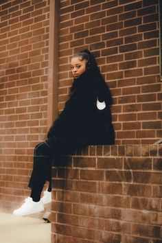 a woman sitting on top of a brick wall next to a tall building and wearing white sneakers