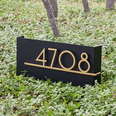 a black and gold address sign sitting in the middle of some grass with trees behind it