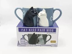 two coffee mugs with cats on them sitting in front of a box that says eat kiss pair